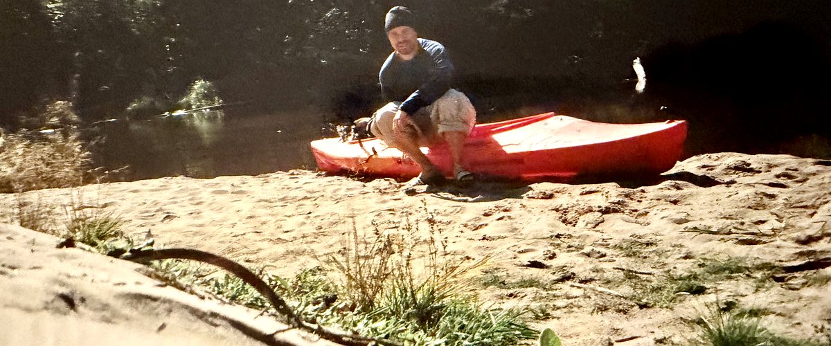 A red kayak pulled up on a sandbank beside the dark, slow water of the Big Two-Hearted River in Michigan's Upper Peninsula