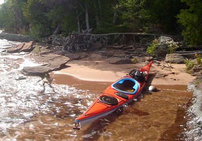 An orange sea kayak resting in shallow water on a sand and sandstone beach along a forested Great Lakes shoreline