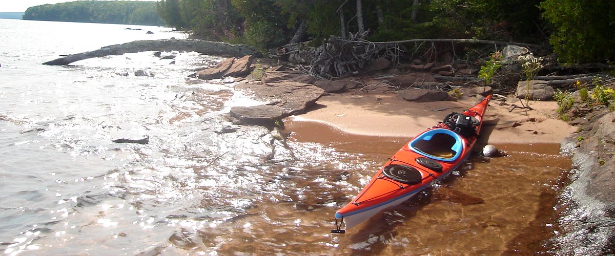 An orange sea kayak resting in shallow water on a sandstone and sand beach along a forested Great Lakes shoreline in Michigan
